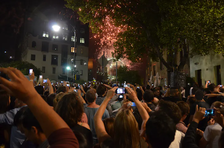A packed crowd takes photos of fireworks visible between buildings.