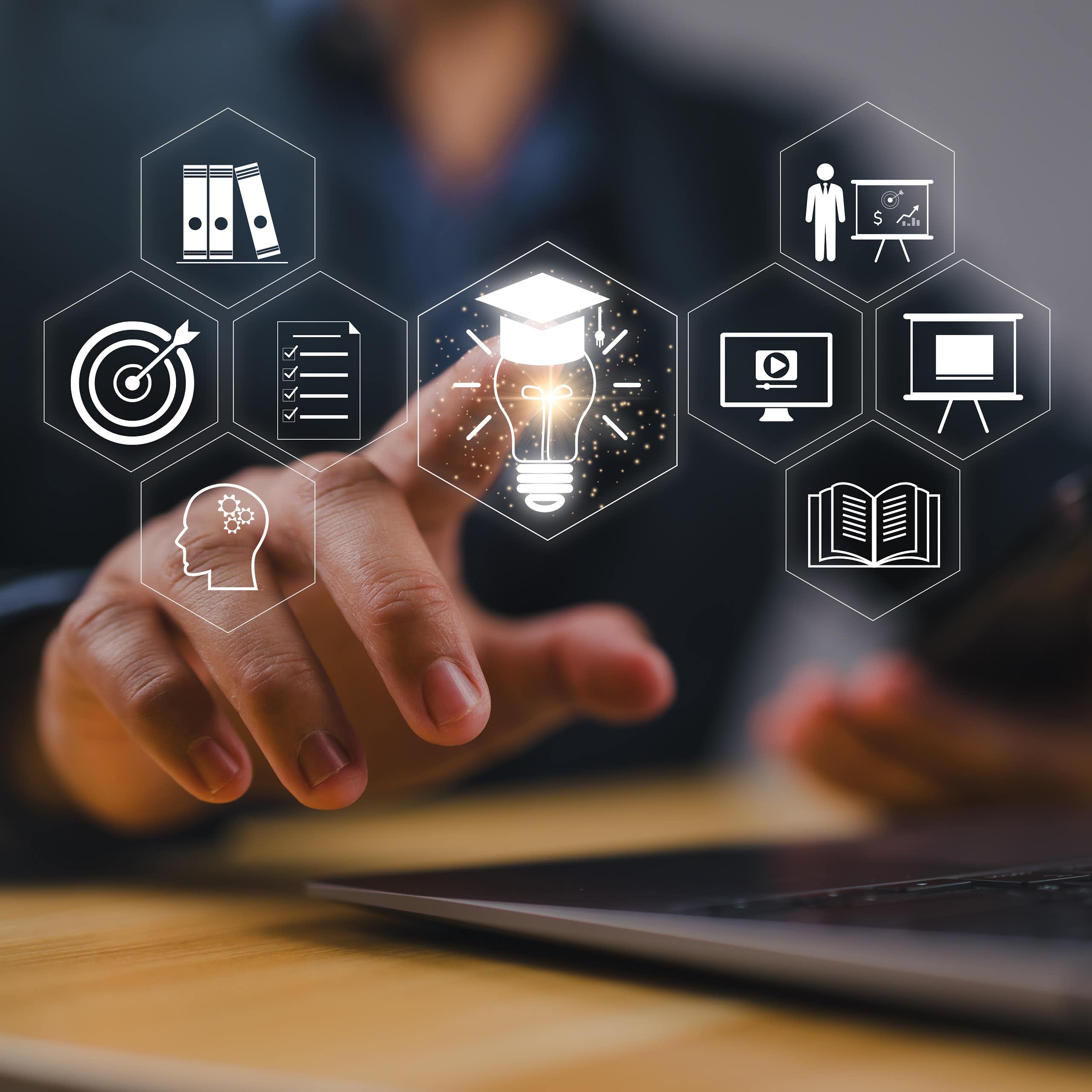A person seated at a table or desk it touching a digital image of a lightbulb with a graduate's cap on top.