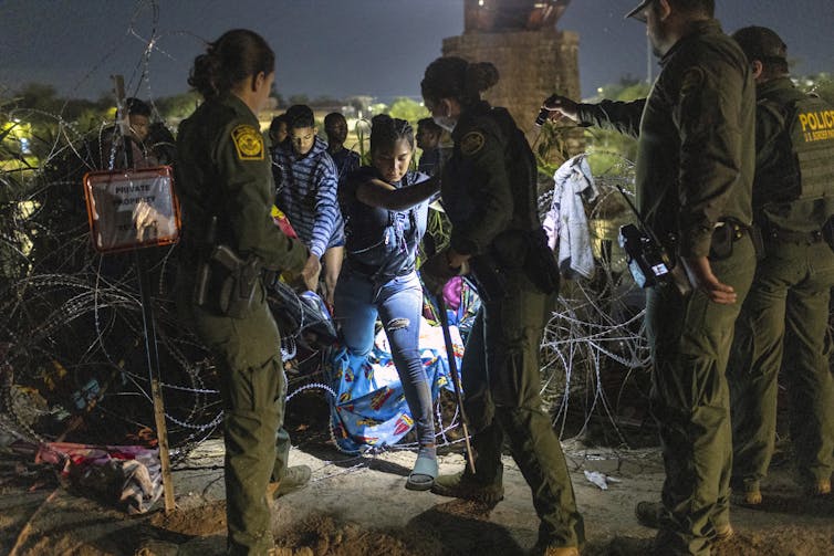 Two women wearing army green uniforms stand near barbed wire and one holds the hand of a woman who steps over the wire. They are there on a dark night.