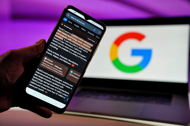 A smartphone help in front of an open laptop displaying the Google logo displays a Google search engine result with a section at the top of the search that says 'AI Overview' followed by an AI-generated response to a search