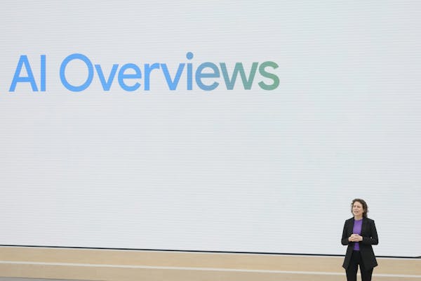 A woman in a suit stands in front of a large screen that displays the phrase 'AI Overviews' on it