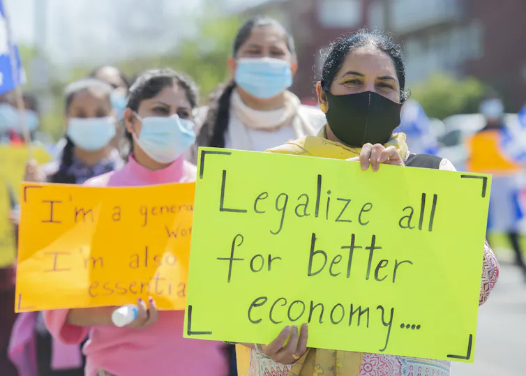 Women wearing face masks carry signs calling for permanent status for migrants, refugees, international students and temporary foreign workers.