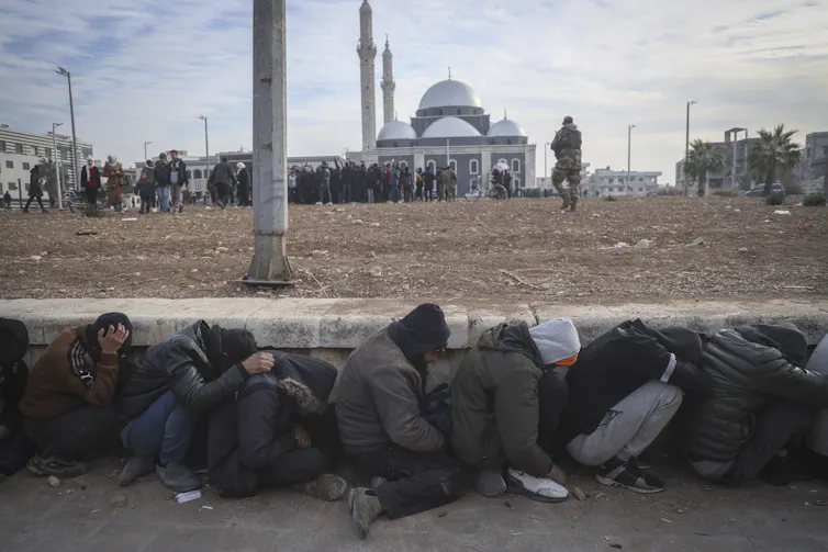People huddle against a retaining wall and hide their heads as an armed fighter walks nearby
