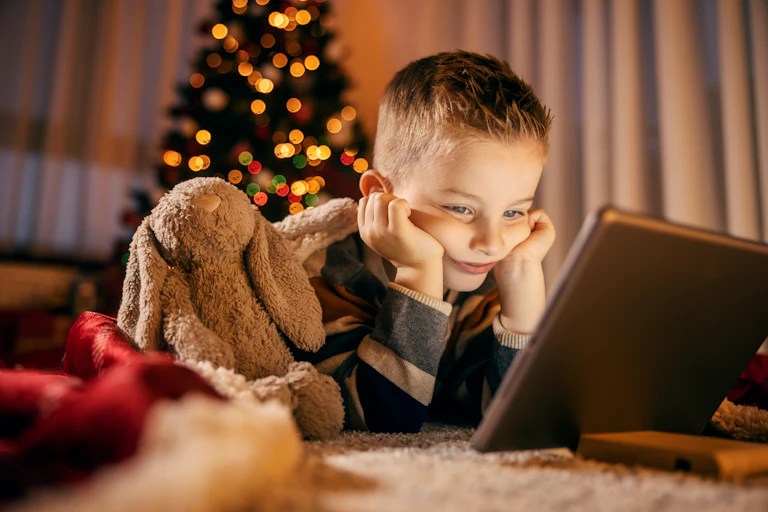 Little boy looks sadly at tablet in front of christmas tree.