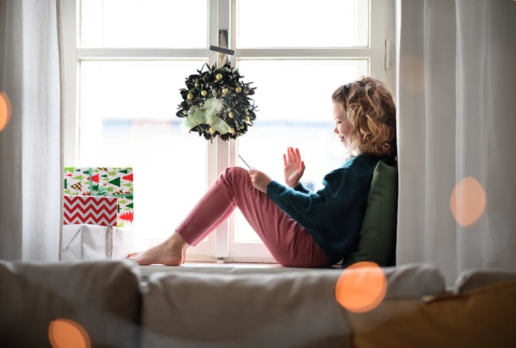 A young teen girl waves at her pone while sitting under a Christmas wreath.