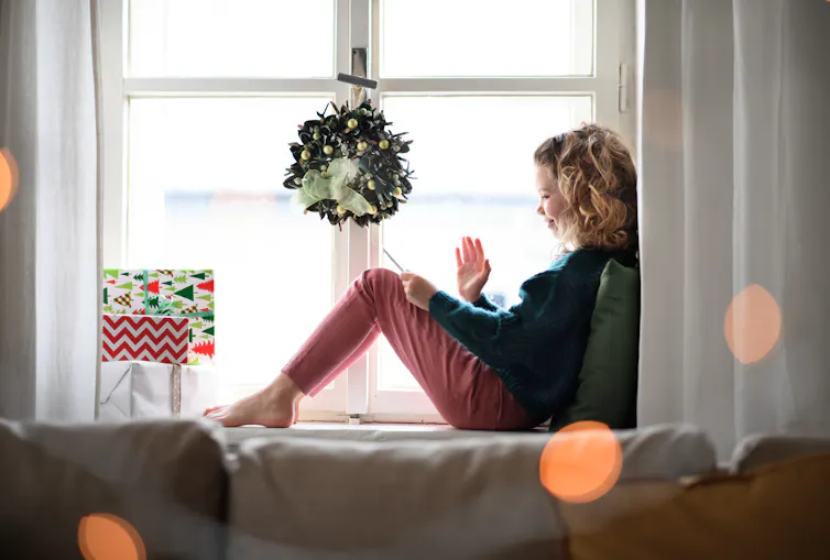 A young teen girl waves at her pone while sitting under a Christmas wreath.
