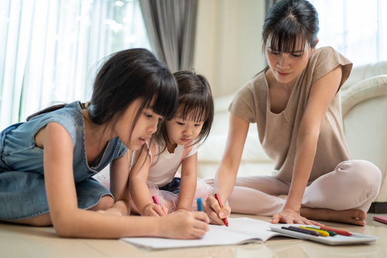 Two girls colour a page on the ground with their mother.