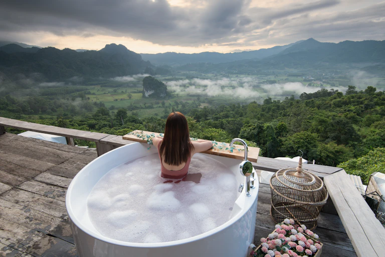 A woman relaxes in a giant outdoor bath while overlooking a green landscape, in a scene depicting luxury travel.
