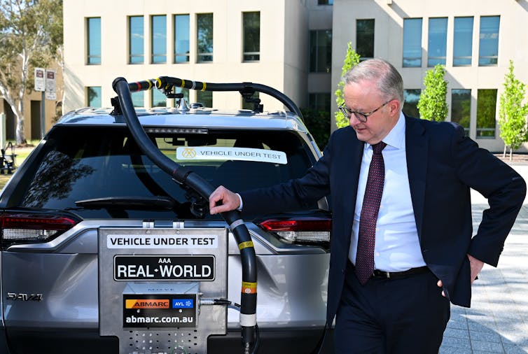 The rear end of a silver car hooked up to a machine testing real world emissions, while Australian Prime Minister Anthony Albanese looks on.