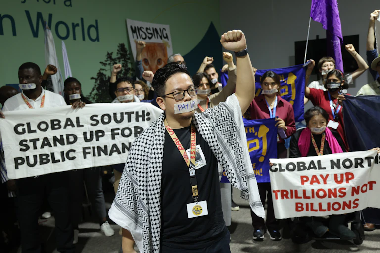 Protestors holding up banners saying 'Global North: pay up trillions not billions' and "Global South: Stand Up for public finance". The protest leader is wearing the Palestinian keffiyeh and their mouth has tape over it.