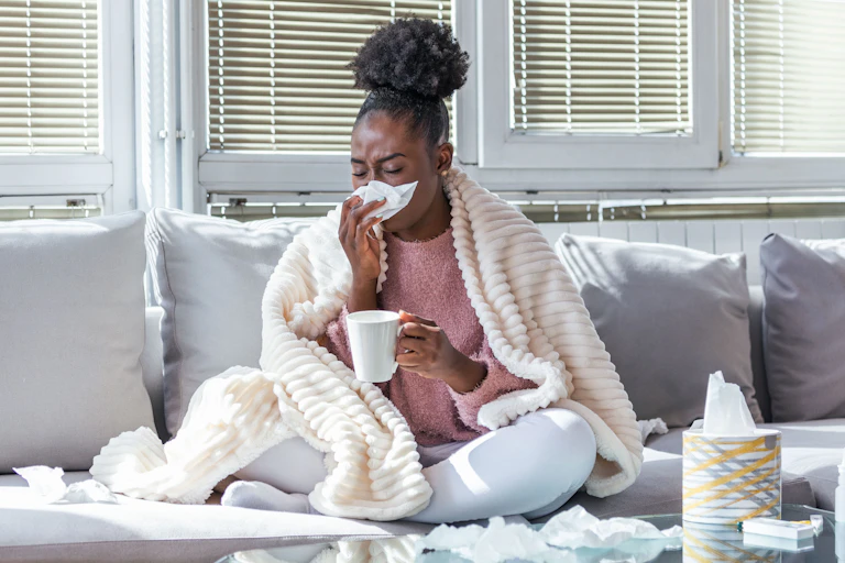 A sick woman sneezes into a tissue while sitting on her couch.