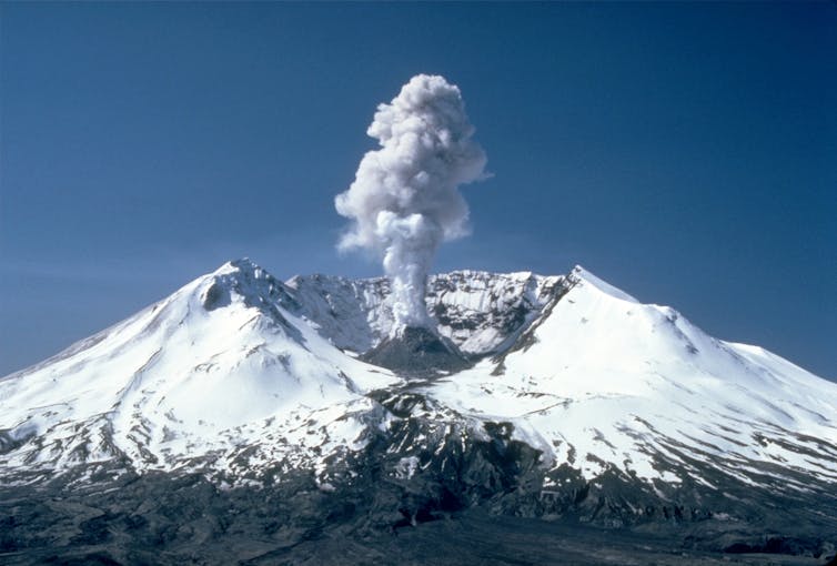 Plumes of steam, gas, and ash coming out of big crater on a conical snow-covered mountain.