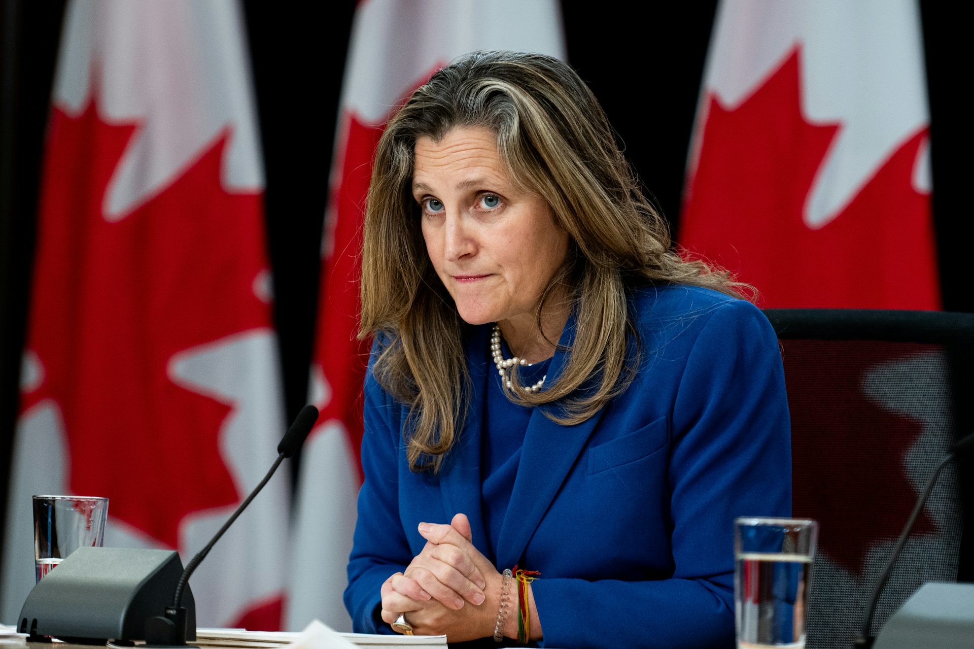 A woman with dark blonde hair in a blue suit with microphone in front of her and Canadian flags behind her.