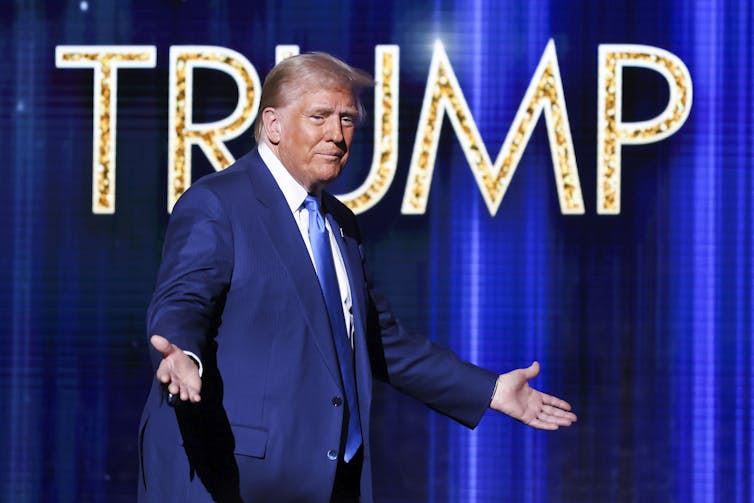 A rotund man in a blue suit with fluffy greyish-blonde hair walks onto a stage with his arms extended. A golden Trump banner is behind him.