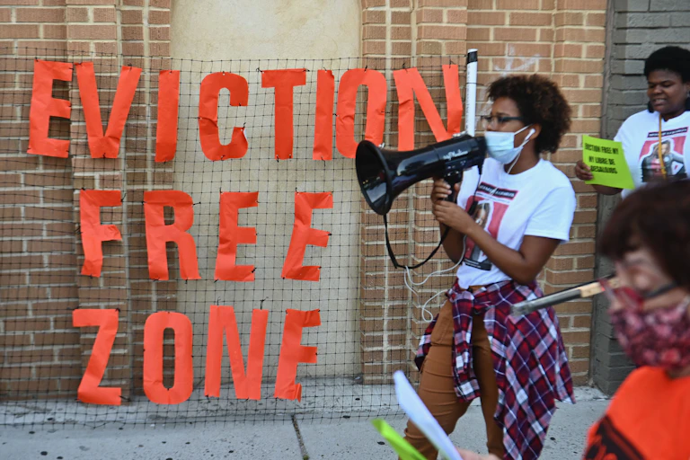 A Black woman with a bullhorn marches by a sign reading "EVICTION FREE ZONE" in red letters