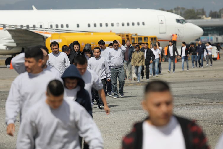 A line of men wearing white walk away from an airplane.