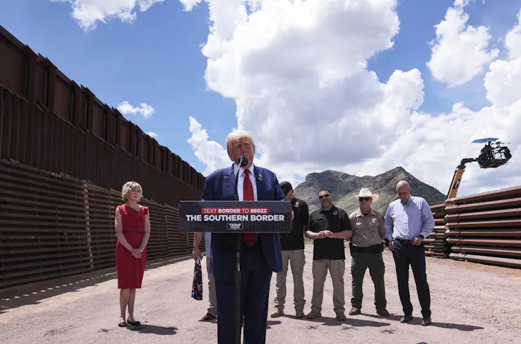 An older man with white hair wears a blue suit and stands at a podium, with several people around him, outside on a clear blue sky day. He stands in front of a very large dark wall.