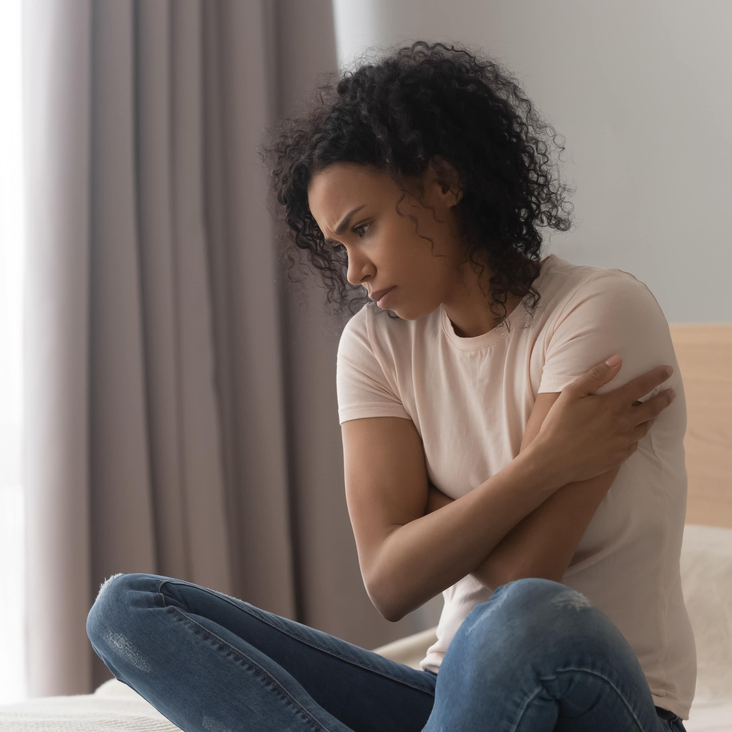 A woman sits cross-legged on her bed with arms crossed and gaze down, appearing sad.