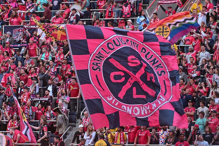 A massive red-and-black flag, at least 12 feet high, waves before crowd of soccer fans. It reads