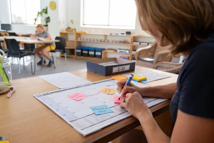 a woman writes on post it notes on a calendar at a teacher's desk in front of a classroom