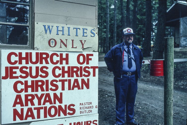 Middle-aged man stands next to two signs, one reading 'Whites Only,' the other reading 'Church of Jesus Christ Christian Aryan Nations.'