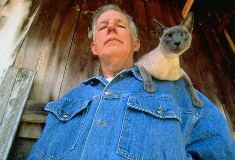 Middle-aged man wearing a denim shirt with a cat perched on his shoulder.