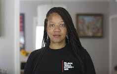 A young Black woman looks into the camera. She is wearing a black sweatshirt with the slogan 'Black Wall Street 100'