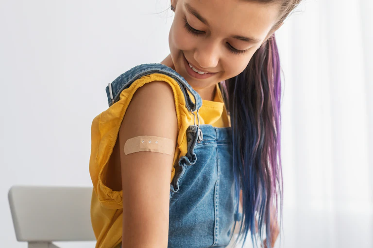 A teenage girl looks down at her upper arm which has a bandaid on.