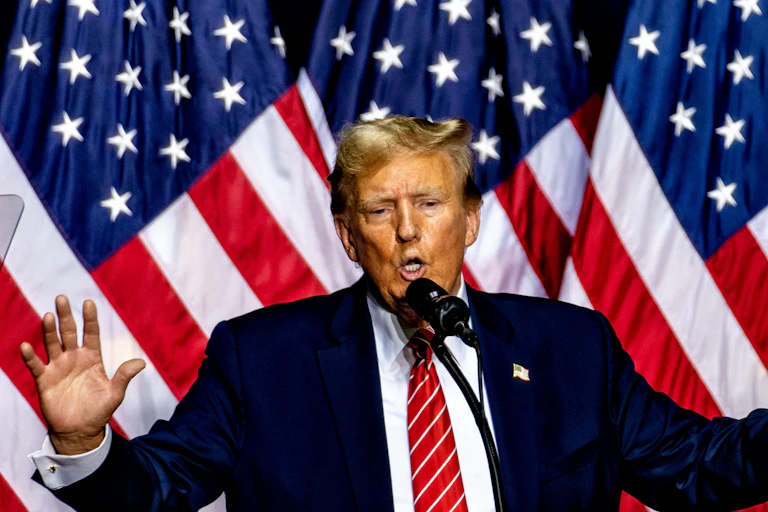 Donald Trump standing in front of three US flags with his arms raised.