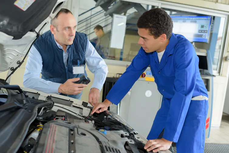 apprentice mechanic looking under a car bonnet with his supervisor