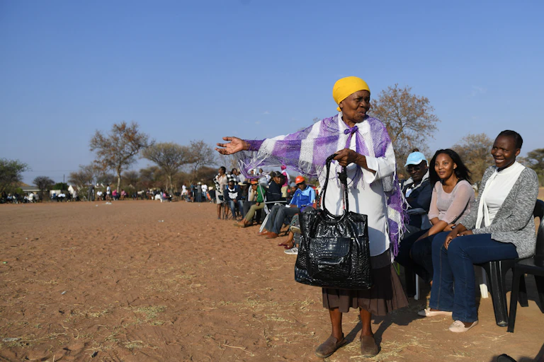 A long queue of seated people on a dusty field. An elderly woman stands in the foreground gesturing and talking.
