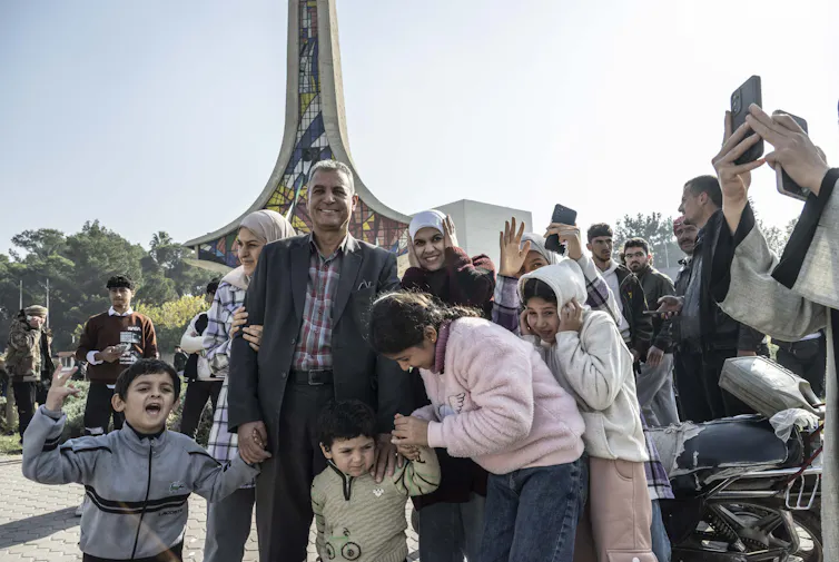 A group with smiling men, women and children gather in a sunny plaza.