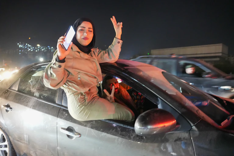 A woman in a tan outfit raising her fingers in a victory sign as she sits in a car window.