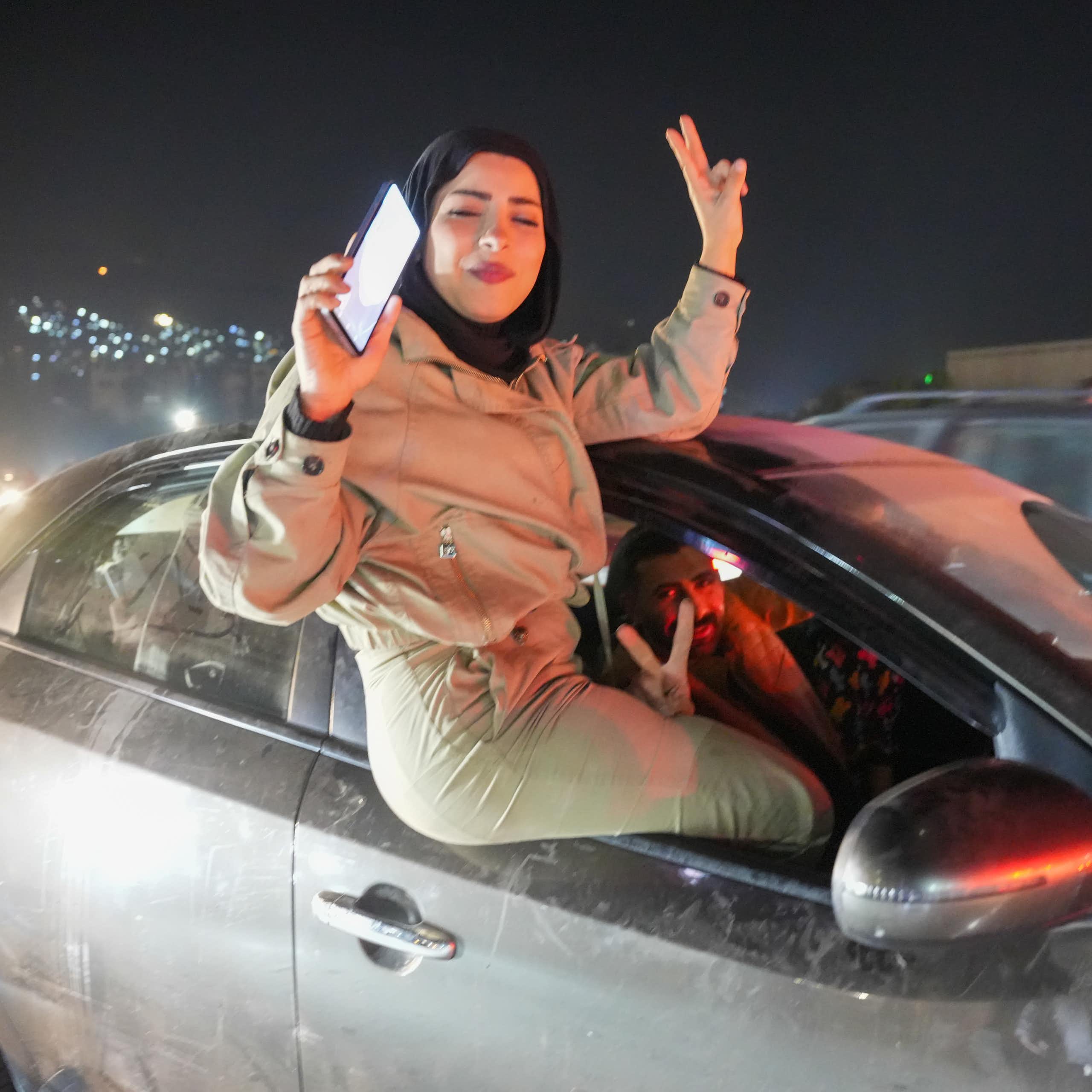 A woman in a tan outfit raising her fingers in a victory sign as she sits in a car window.