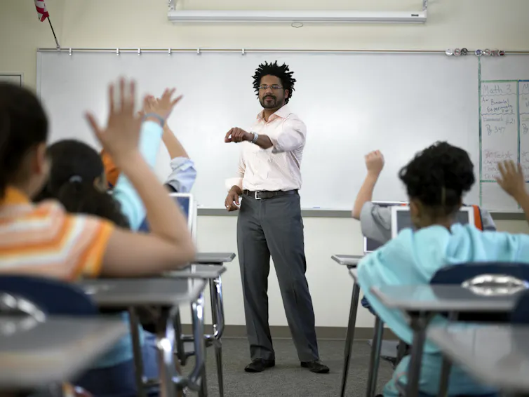 Black male teacher stands in front of classroom and points to student with hand raised