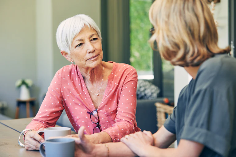 A woman with short, white hair, who is wearing a pink shirt, sits and looks thoughtfully at another woman who appears to be talking.