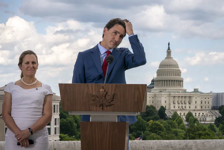 A dark-haired man runs his fingers through his hair as a small woman in a white dress stands behind him. In the background is the U.S. Capitol building.