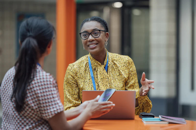 a black woman wearing glasses and a yellow shirt sits in a front of a desk with a computer gesturing across from another woman