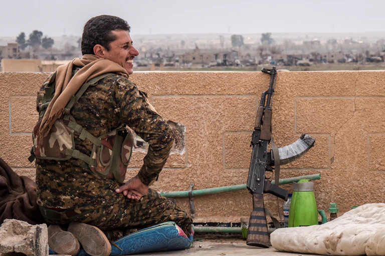 A soldier in position on top of a building with his gun resting on the wall.
