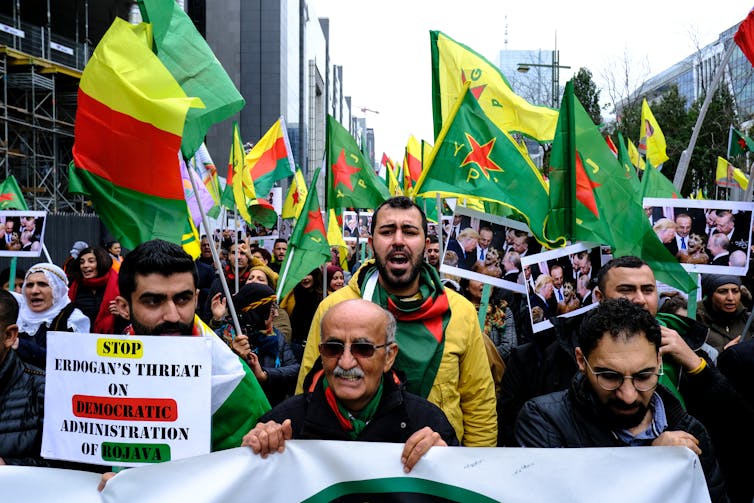 A crowd of people with flags and a sign reading 'stop Erdogan's threat on democratic administration of Rojava'.
