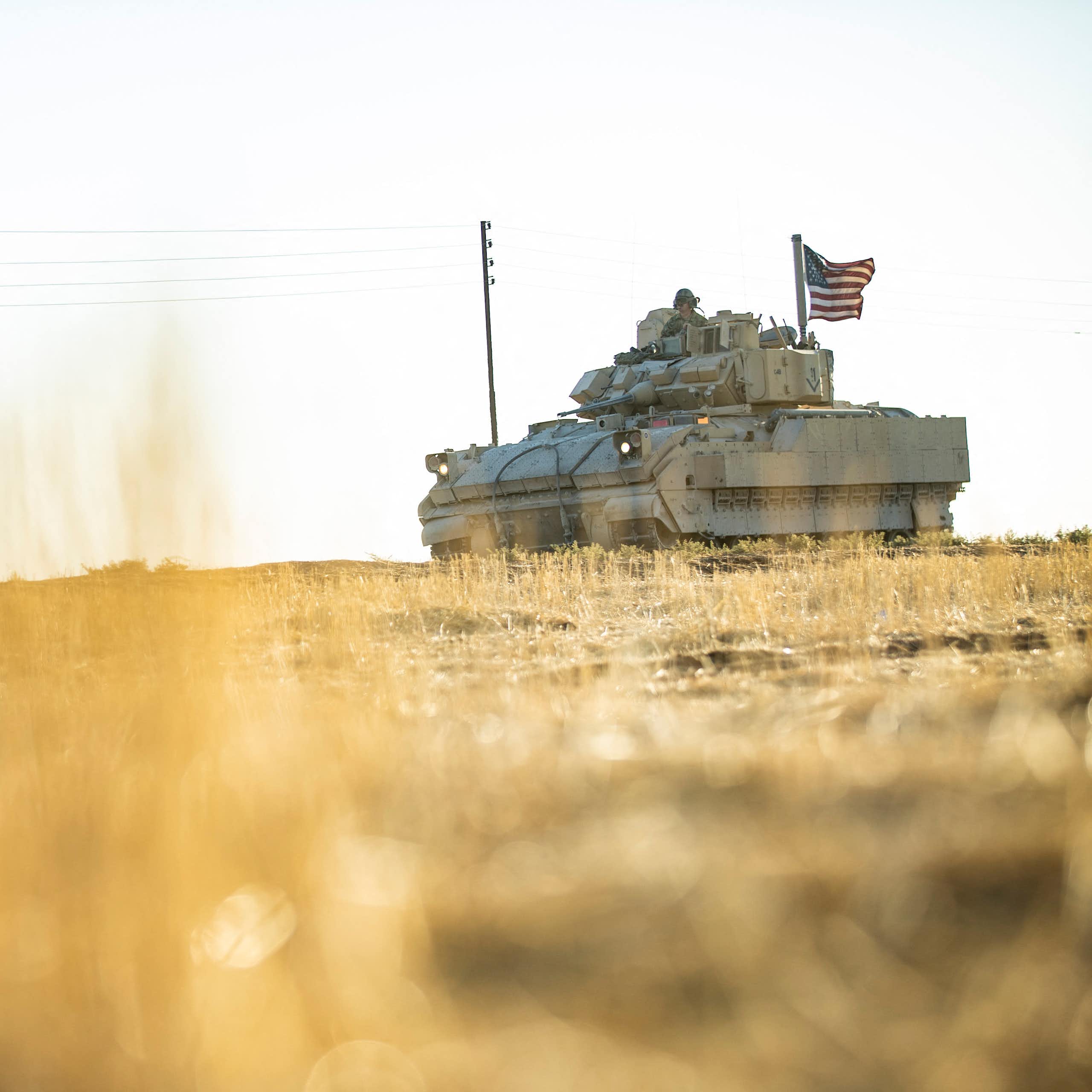 A grey tank is seen on a field against a grey sky. The tank has a US flag flying from it.