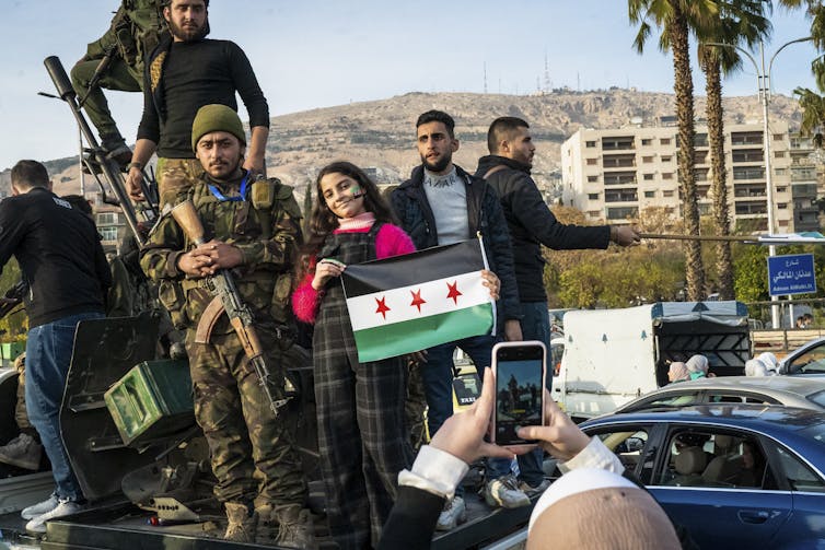 A girl stands and holds a black and green and white flag with three red stars and stands next to several men, including one in camouflage holding a gun.
