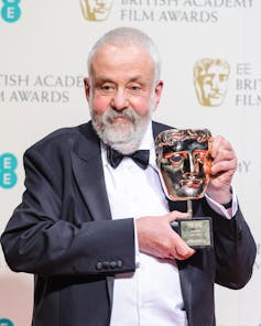 An older man with grey hair and beard dressed in a tuxedo holding up a Bafta award.