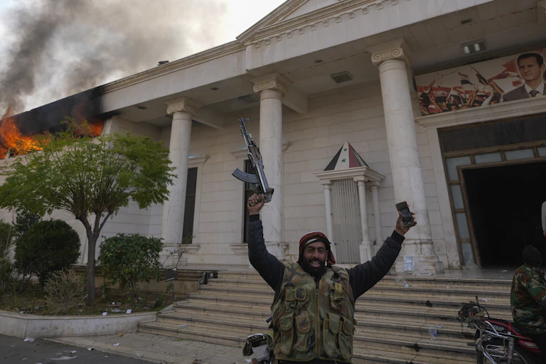 An armed man smiles with his arms raised in front of a burning court building.