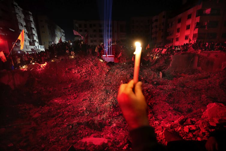 A hand holding up a candle in front of a pile of rubble illuminated by red lights.