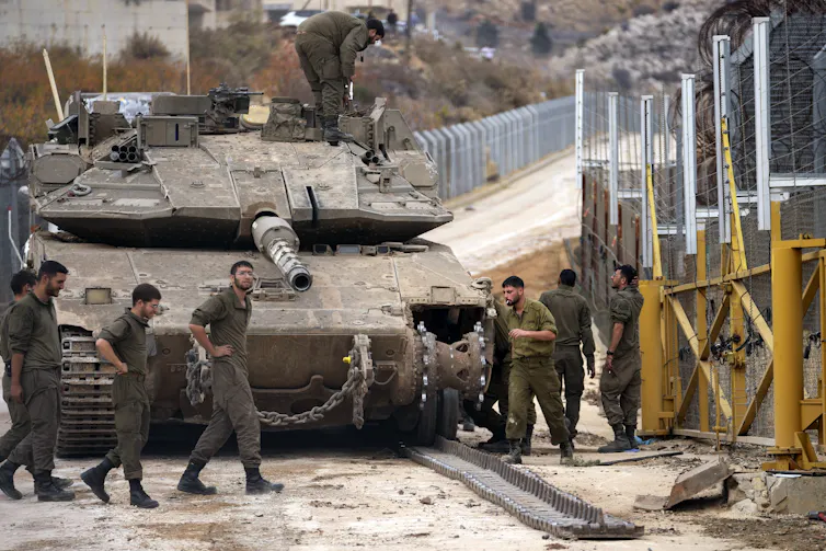 Soldiers stand around a tank next to a fortified fence.