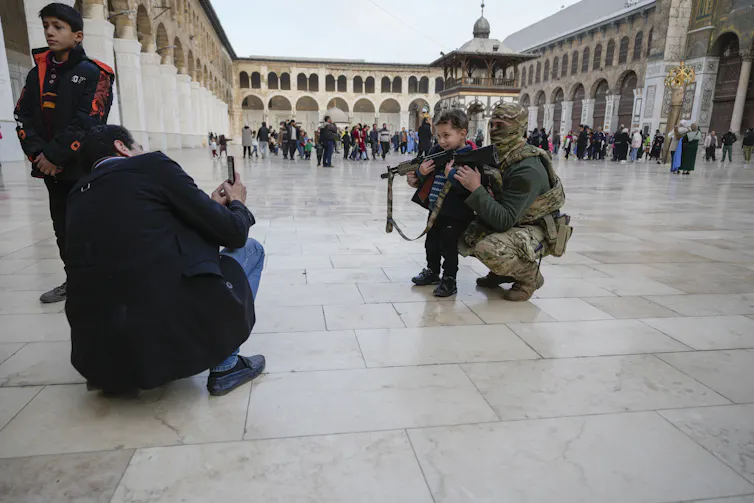 A small boy poses for a photo with a heavily armed fighter with a camouflaged scarf around in his an outdoor courtyard.