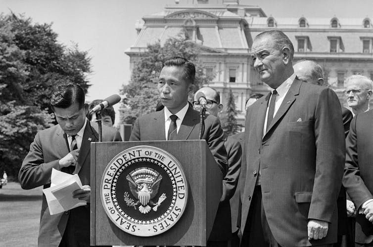 Two men in suits stand behind a platform.