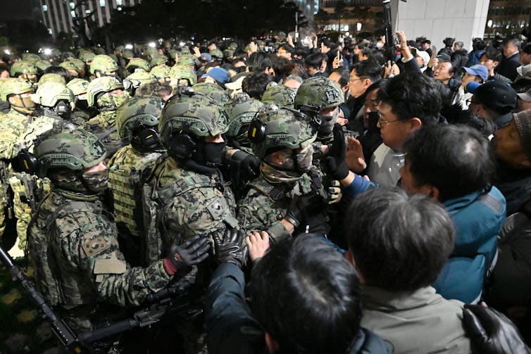 Soldiers in riot gear confront angry looking protesters.