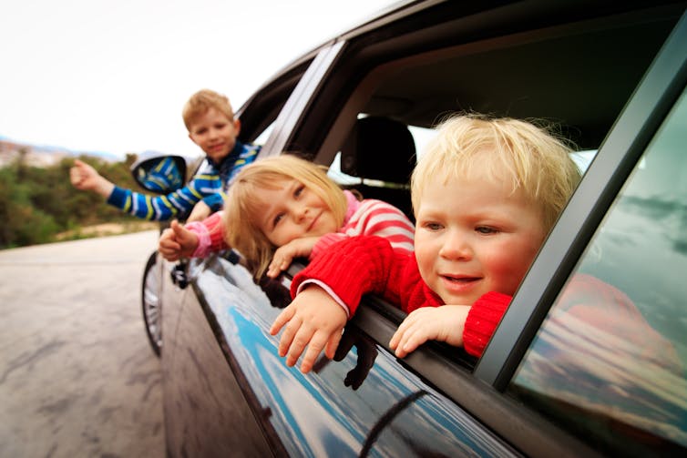 Three young kids hang out the window of a car.